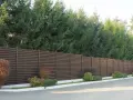 Nowoczesny, brązowy płot lamelowy to świetny sposób, by czym zasłonić płot od sąsiada. Za nim rosną wysokie świerki.