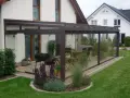 Nowoczesny ogród zimowy z meblami i grillem, otoczony zielonym trawnikiem i roślinnością.