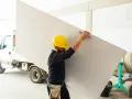 Budowlaniec w żółtym kasku niesie płytę gipsowo-kartonową, która posłuży do budowy ścianki działowej z gk.