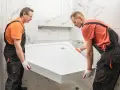 Dwóch mężczyzn w roboczych kombinezonach przenosi biały brodzik prysznicowy. Pokazują, jak zamontować brodzik na styropianie.