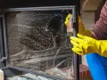 Ręce w żółtych rękawiczkach czyszczą zaplamioną szybę kominka. Dowiedz się, jak wyczyścić kominek w środku, aby znów lśnił.