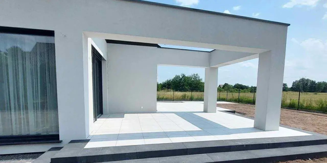 Nowe, białe płytki tarasowe lśnią na tle nowoczesnej architektury. Trwają prace nad układaniem płyt tarasowych, tworząc elegancką przestrzeń.