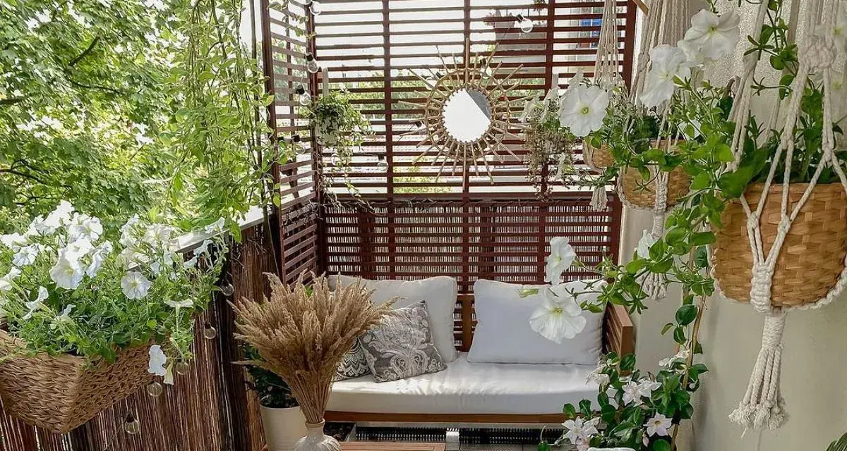 Przytulny balkon z drewnianą sofą, białymi kwiatami i ozdobnym lustrem w kształcie słońca.