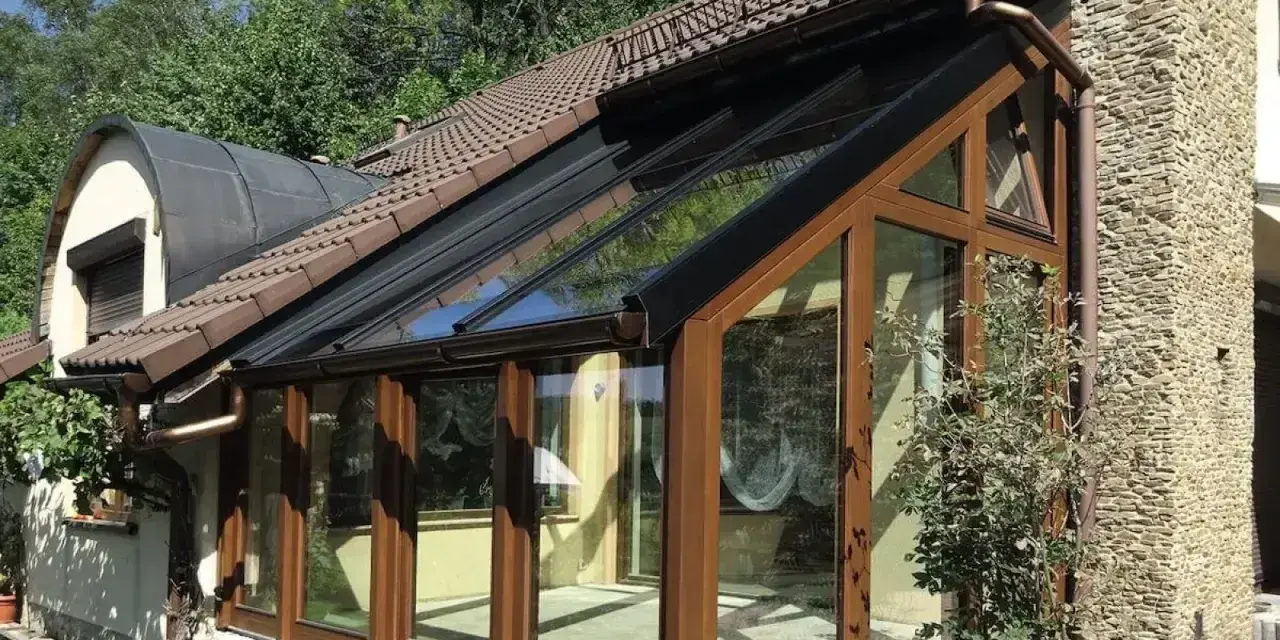 Nowoczesna budowa ogrodu zimowego z drewnianymi ramami i przeszklonym dachem, przylegająca do kamiennego domu.