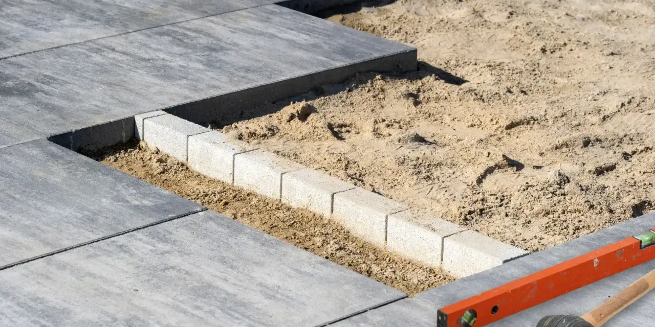 Układanie płyt tarasowych na podsypce piaskowej. Widać fragmenty gotowego tarasu, podsypkę i obrzeże z kostki. Narzędzia budowlane leżą obok.