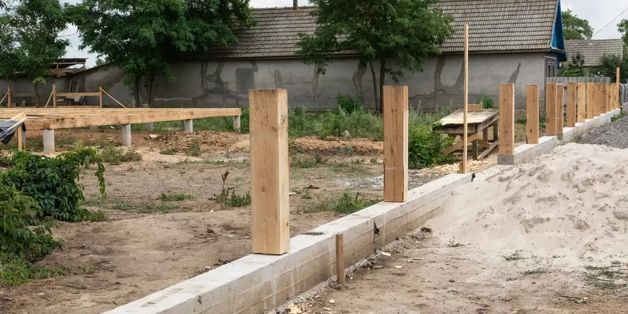 Rozpoczyna się montaż ogrodzenia. Widać betonowy fundament, drewniane słupki i budowę podłogi na podwyższeniu.