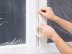 Ręce przyklejają taśmę malarską, by zabezpieczyć okna przed malowaniem. Folia chroni szyby i ramy.