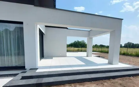 Nowe, białe płytki tarasowe lśnią na tle nowoczesnej architektury. Trwają prace nad układaniem płyt tarasowych, tworząc elegancką przestrzeń.