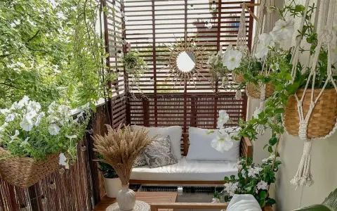 Przytulny balkon z drewnianą sofą, białymi kwiatami i ozdobnym lustrem w kształcie słońca.