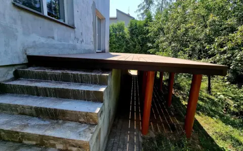 Nowy, wysoki taras na słupach, z drewnianym blatem, sąsiaduje z betonowymi schodami i zielonym ogrodem.