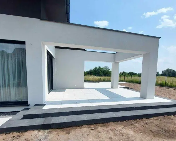 Nowe, białe płytki tarasowe lśnią na tle nowoczesnej architektury. Trwają prace nad układaniem płyt tarasowych, tworząc elegancką przestrzeń.