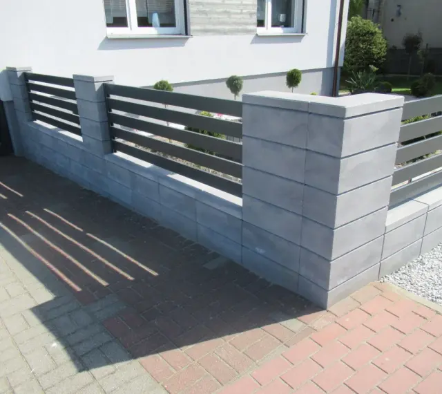 Nowoczesne ogrodzenie z betonowych słupków i ciemnoszarych paneli. Kolor ogrodzenia idealnie komponuje się z elewacją domu.