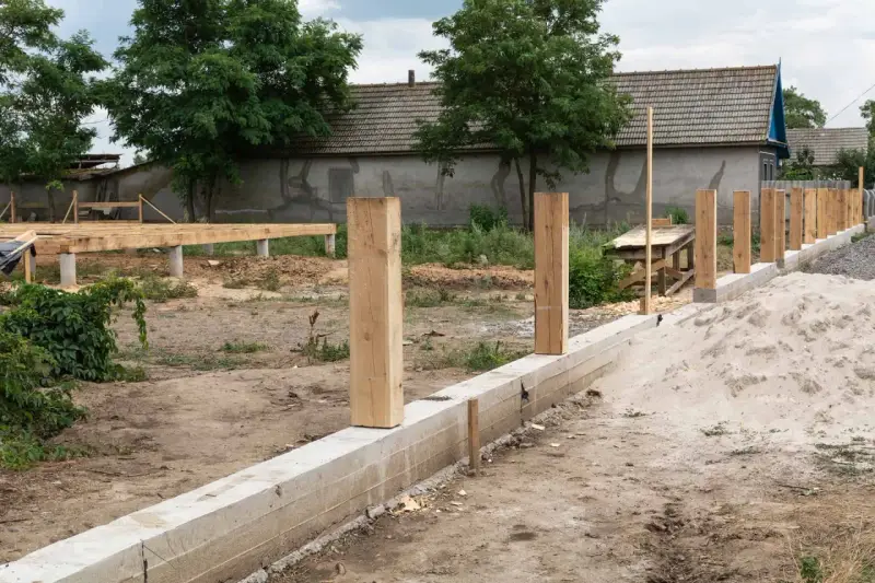 Rozpoczyna się montaż ogrodzenia. Widać betonowy fundament, drewniane słupki i budowę podłogi na podwyższeniu.